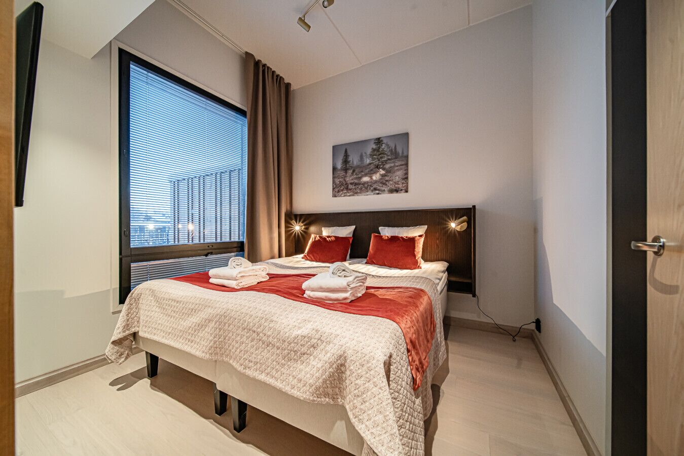 Ein modernes Hotelzimmer mit einem großen Bett, einem an der Wand befestigten Kopfteil mit integrierter Beleuchtung, einem Fenster mit Jalousien und einem Gemälde.
