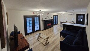 Un salon moderne avec un téléviseur, un canapé et une table basse en verre. On y trouve une cheminée avec un détail rouge, ainsi qu’un coin repas avec un bar.