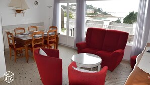 Een kamer met een eettafel, stoelen, een rode bank, een kleine witte tafel en uitzicht op de kust.