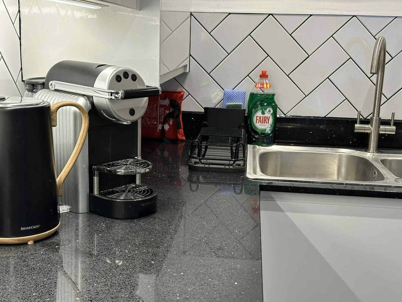 A kitchen with a coffee maker, a kettle, a dish rack, a sink, and a bottle of dish soap.