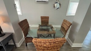 A dining area with a glass-top table surrounded by wicker chairs, an air conditioning unit, and a decorative mirror on the wall. - New -1st Floor Spacious Kit W&D Close 2 Everything 3Mi To Beach Sleeps 4 KingBed (Bonita Springs)