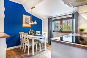 A modern kitchen with a blue accent wall, a dining area with white furniture, and a large window with a view of the outside. - Apartment '2' with Modern Country-Style Kitchen (Cuxhaven)
