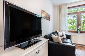 A modern living room with a flat-screen TV on a stand, a black leather sofa with decorative pillows, and a window with a view of greenery. - Apartment '2' with Modern Country-Style Kitchen (Cuxhaven)