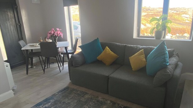 Ein Wohnzimmer mit grauem Sofa, einem Couchtisch und einem Esstisch mit Stühlen. Ein großes Fenster bietet Blick ins Grüne.