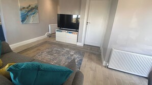 Ein Wohnzimmer mit Sofa, einem türkisfarbenen Kissen, einem Gemälde, einem Fernseher und einer weißen Tür.