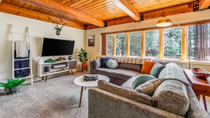 A cozy living room with a wooden ceiling, a large window with a view of trees, a flat-screen TV, a sofa with cushions, a coffee table, and a white entertainment unit. - Cub Cabin Escape: Hot Tub, Games, Fire Pit, BBQ (Big Bear)