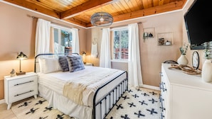 A bedroom with a wooden ceiling, a bed with white bedding, a nightstand with a lamp, and a dresser with decorative items. - Cub Cabin Escape: Hot Tub, Games, Fire Pit, BBQ (Big Bear)