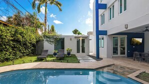 Un espace piscine moderne comprenant une piscine à eau claire, une petite pelouse et une terrasse aménagée avec du mobilier d’extérieur.