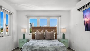 Une chambre à coucher avec un lit en bois, deux tables de chevet avec des lampes, une grande fenêtre donnant sur des immeubles et un téléviseur fixé au mur.
