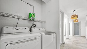 Une buanderie équipée de deux machines à laver blanches, d’une étagère où sont rangées des bouteilles de lessive, et d’un couloir menant à une porte.