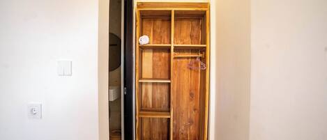 A wooden wardrobe with multiple shelves and hanging space, located in a hallway with a white wall and a door leading to another room.