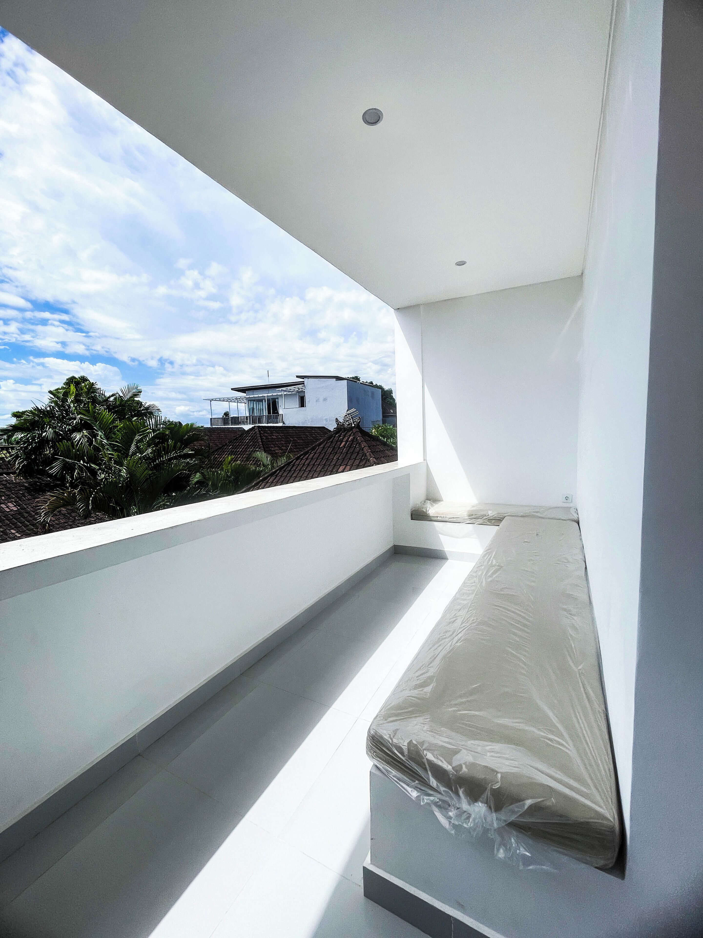 Una habitación moderna y minimalista con un ventanal grande que da a una zona residencial. La habitación tiene un techo blanco, una cama pequeña con colchón y una pared blanca.