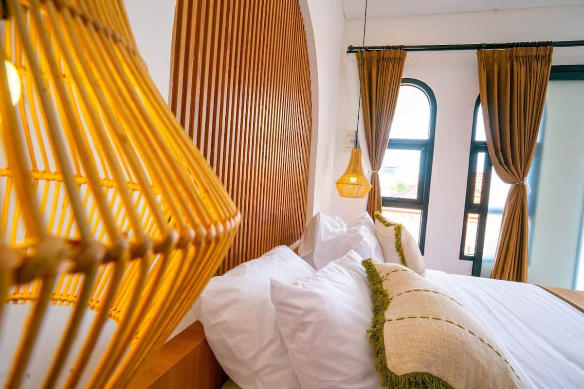 Ein Bett mit weißer Bettwäsche, ein gelber gewebter Sessel und ein Fenster mit Vorhängen.