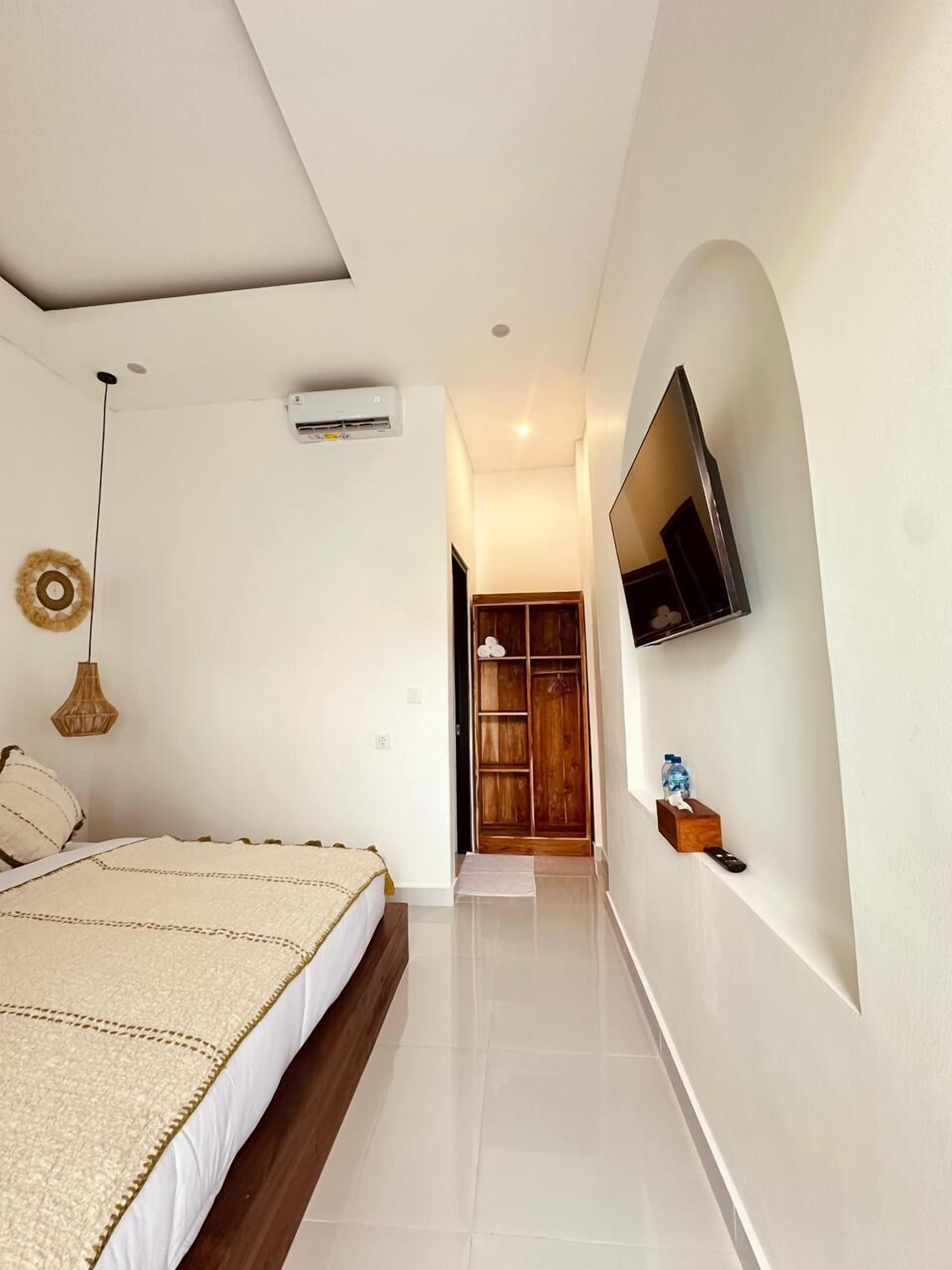 Une chambre à coucher avec un lit, un téléviseur fixé au mur, une armoire en bois et une petite table sur laquelle se trouvent une bouteille et des mouchoirs.