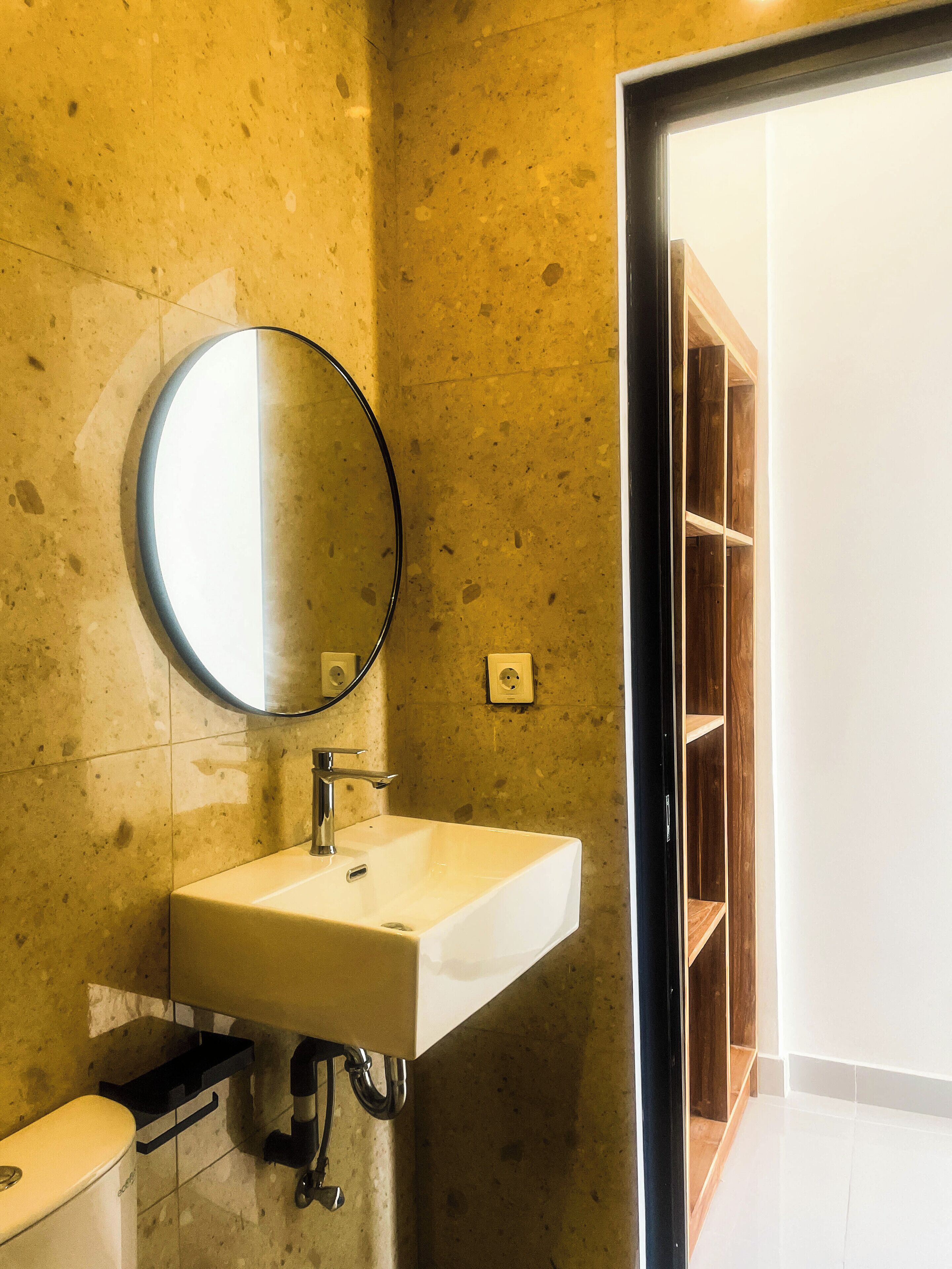 A bathroom with a white sink, a round mirror, and a wooden shelving unit.