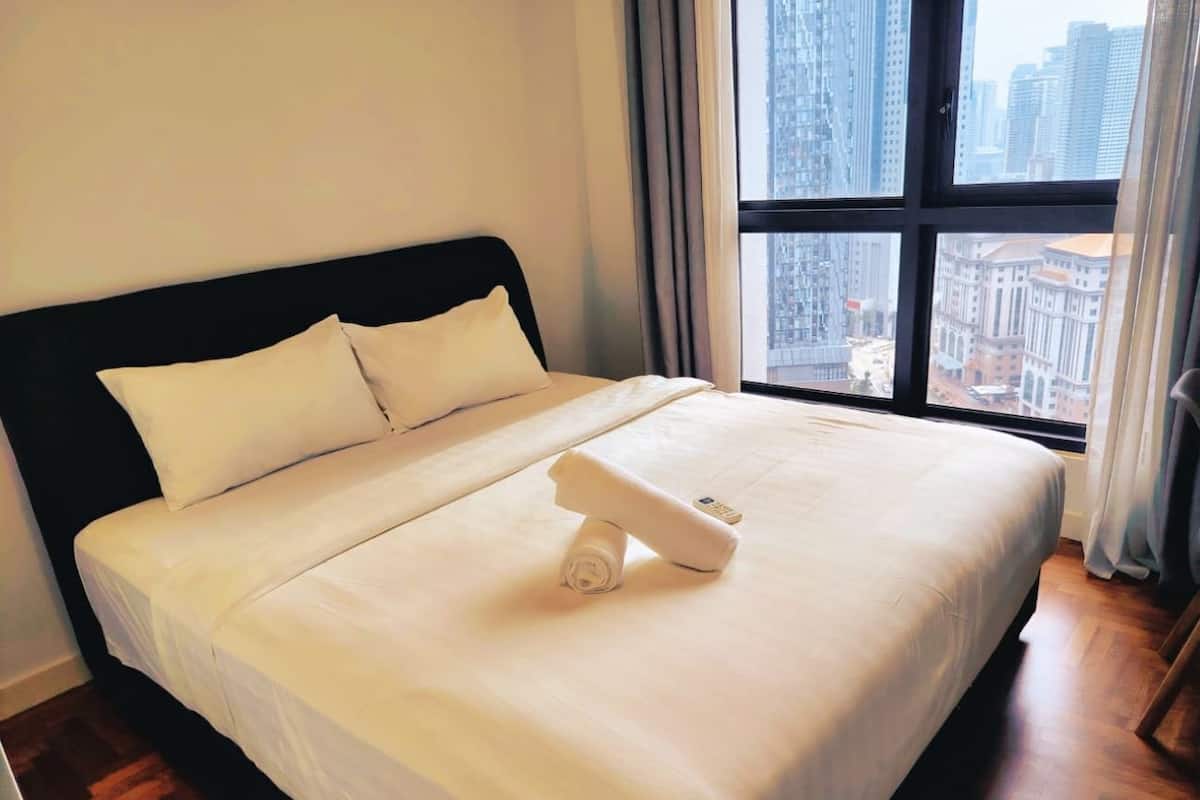 A neatly made bed with a remote control and a rolled towel, a large window with a city view, and wooden flooring.