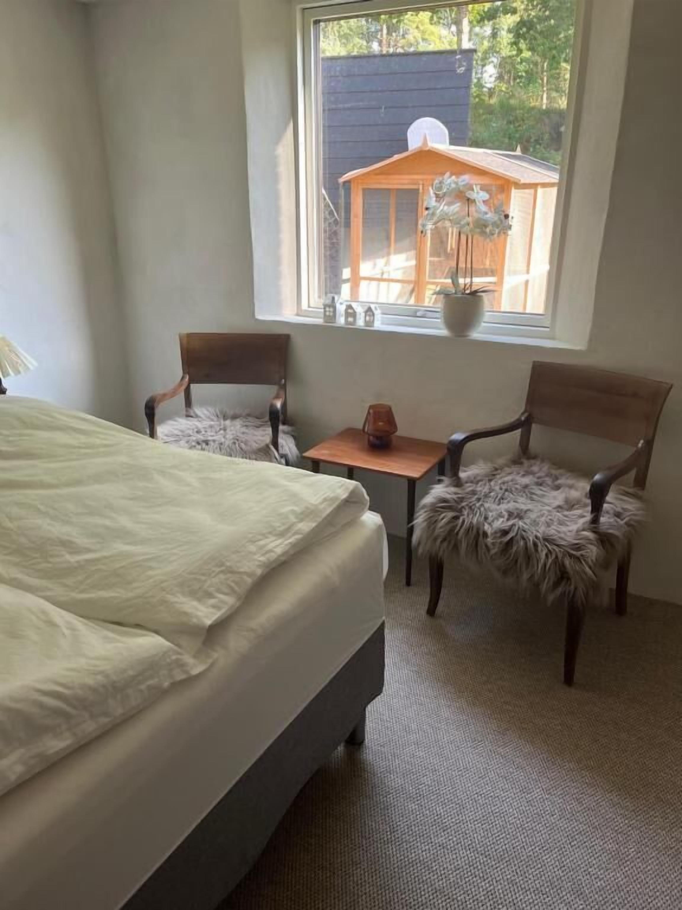 A bedroom with a bed, two chairs, a small table, and a window with a view of a wooden structure outside.