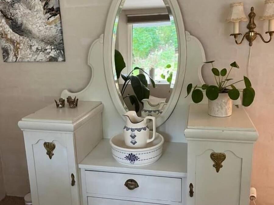 A white vanity with an ornate mirror, a small cabinet with drawers, and a decorative pitcher on top.