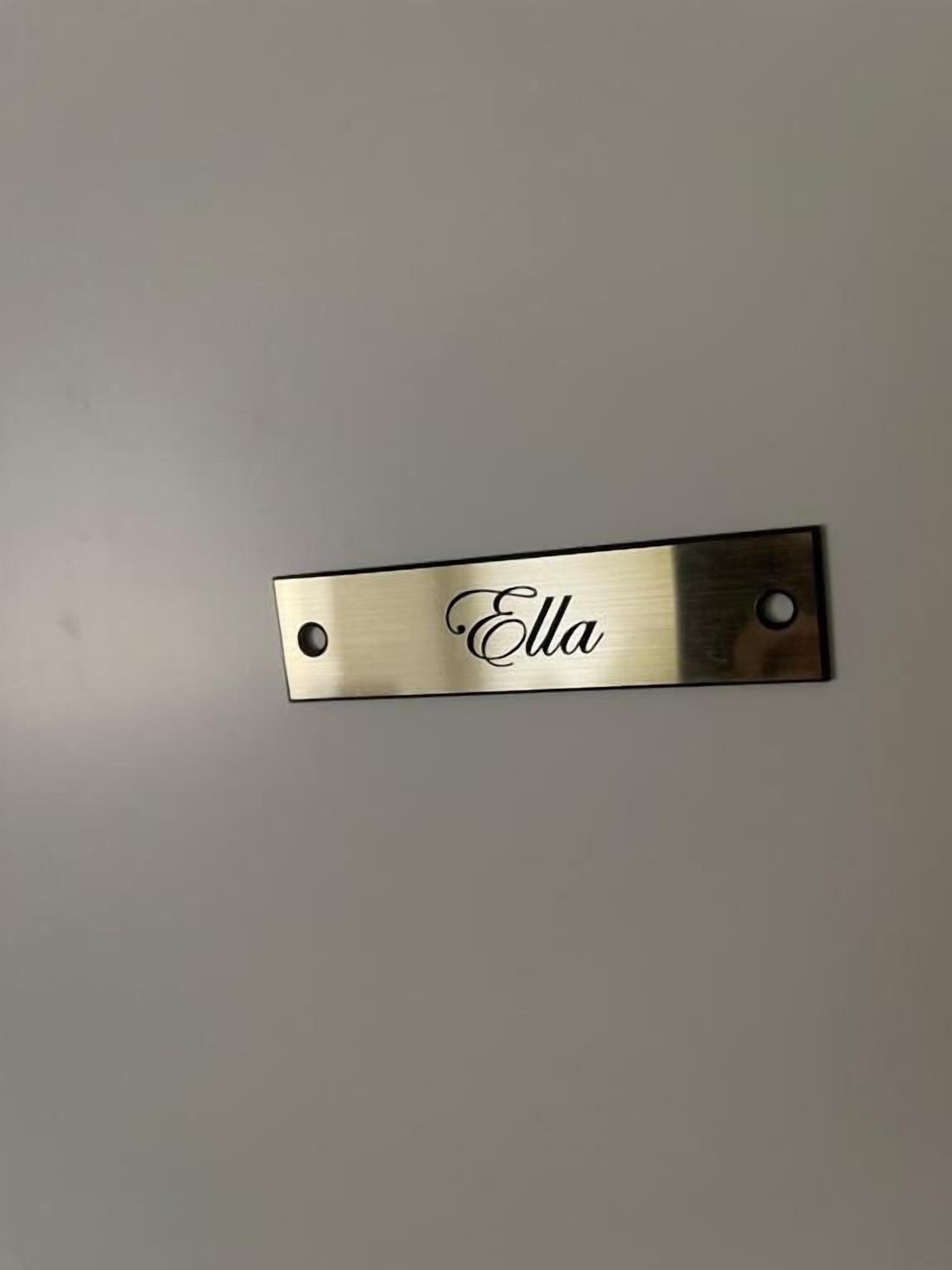 A metallic nameplate with the name "Ella" on a plain wall.