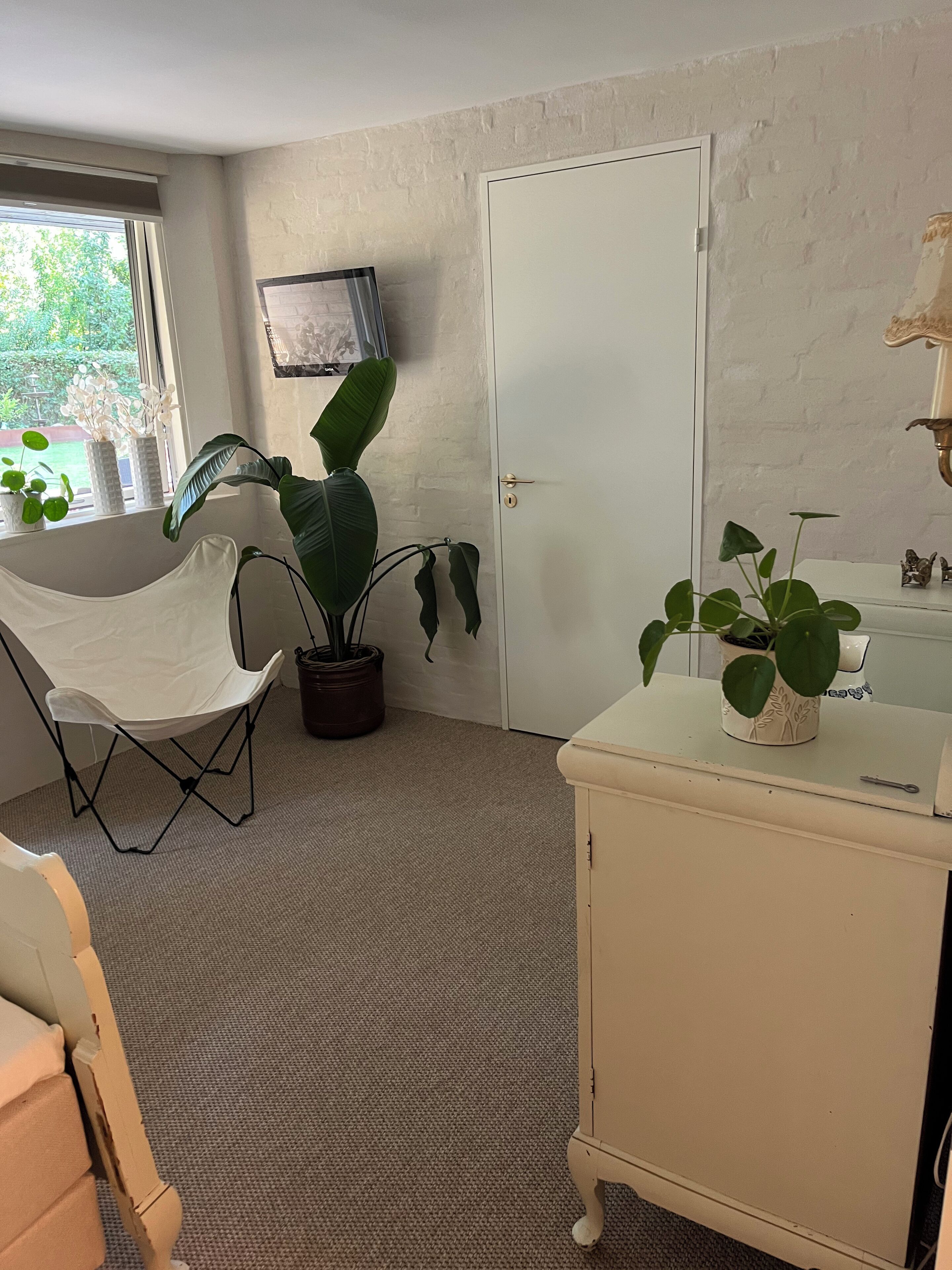 A room with a white door, a window with a view of greenery, a white chair, a potted plant, and a small cabinet with a plant on top.