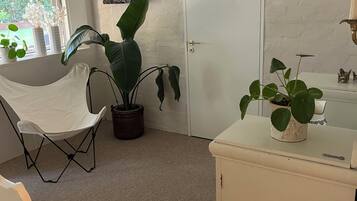 A room with a white door, a window with a view of greenery, a white chair, a potted plant, and a small cabinet with a plant on top.
