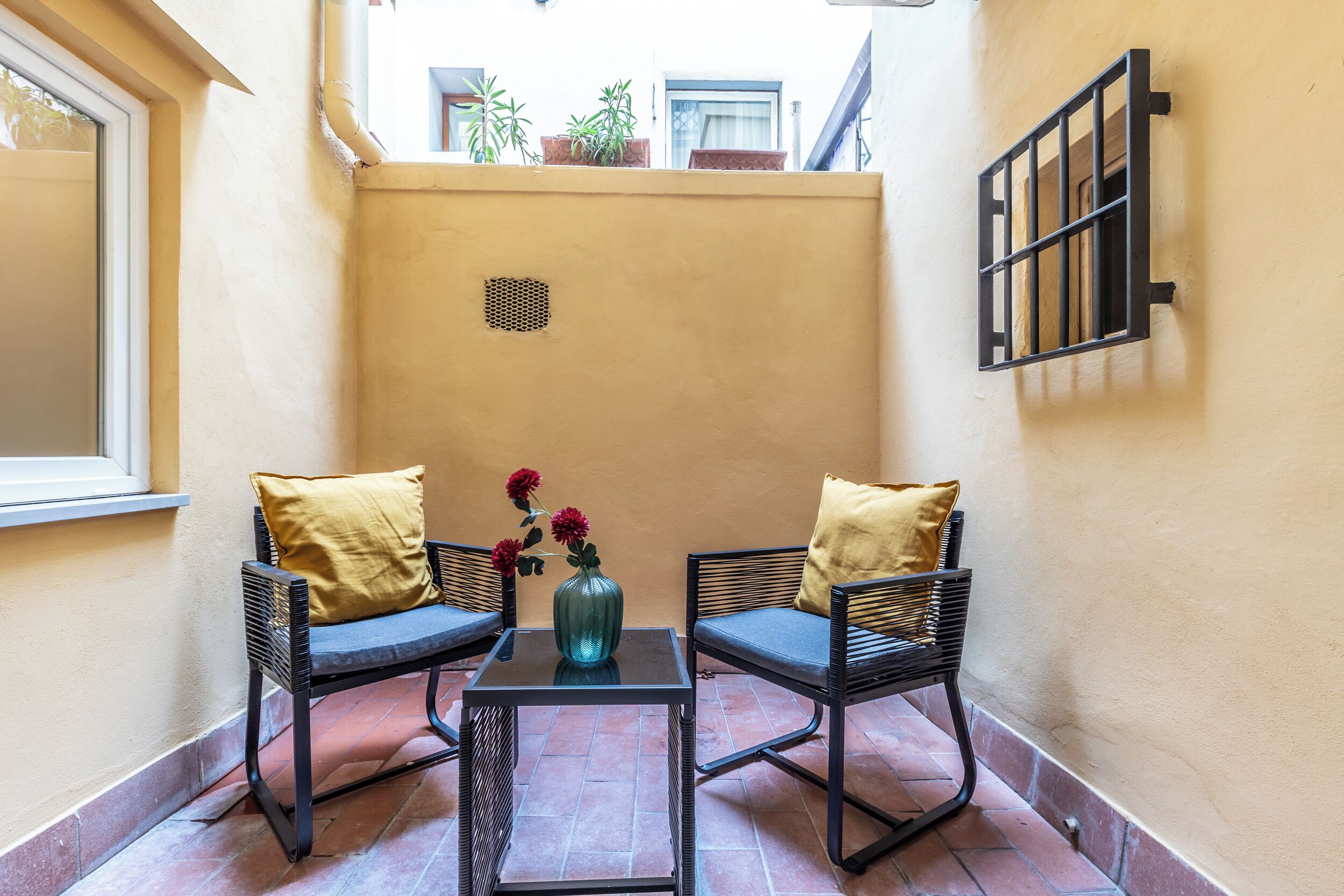 Un pequeño patio con dos sillas de mimbre, una mesa de vidrio y un florero con flores.