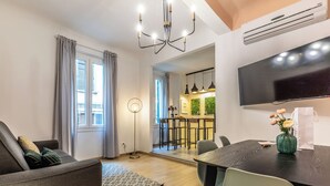 A modern living room with a sofa, dining table, and chairs. There is a TV mounted on the wall, a kitchen area visible through an open door, and a large window with curtains. - Wine Apartments Florence Biancone (Florence)