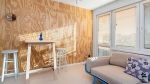 Ein Zimmer mit einer Holzwand, einem weißen Tisch mit zwei Hocker, einem beigefarbenen Sofa mit einem gemusterten Kissen und einem Fenster mit Jalousien.