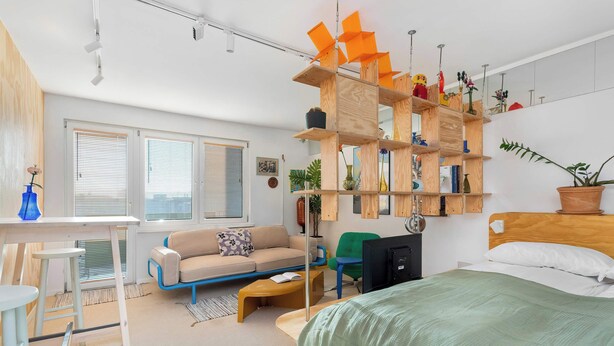 Ein modernes, gut beleuchtetes Wohnzimmer mit einer Couch, einem Couchtisch und einem Bett. Regale mit Pflanzen und Dekoration sowie ein Schreibtisch mit Stuhl sind ebenfalls vorhanden.