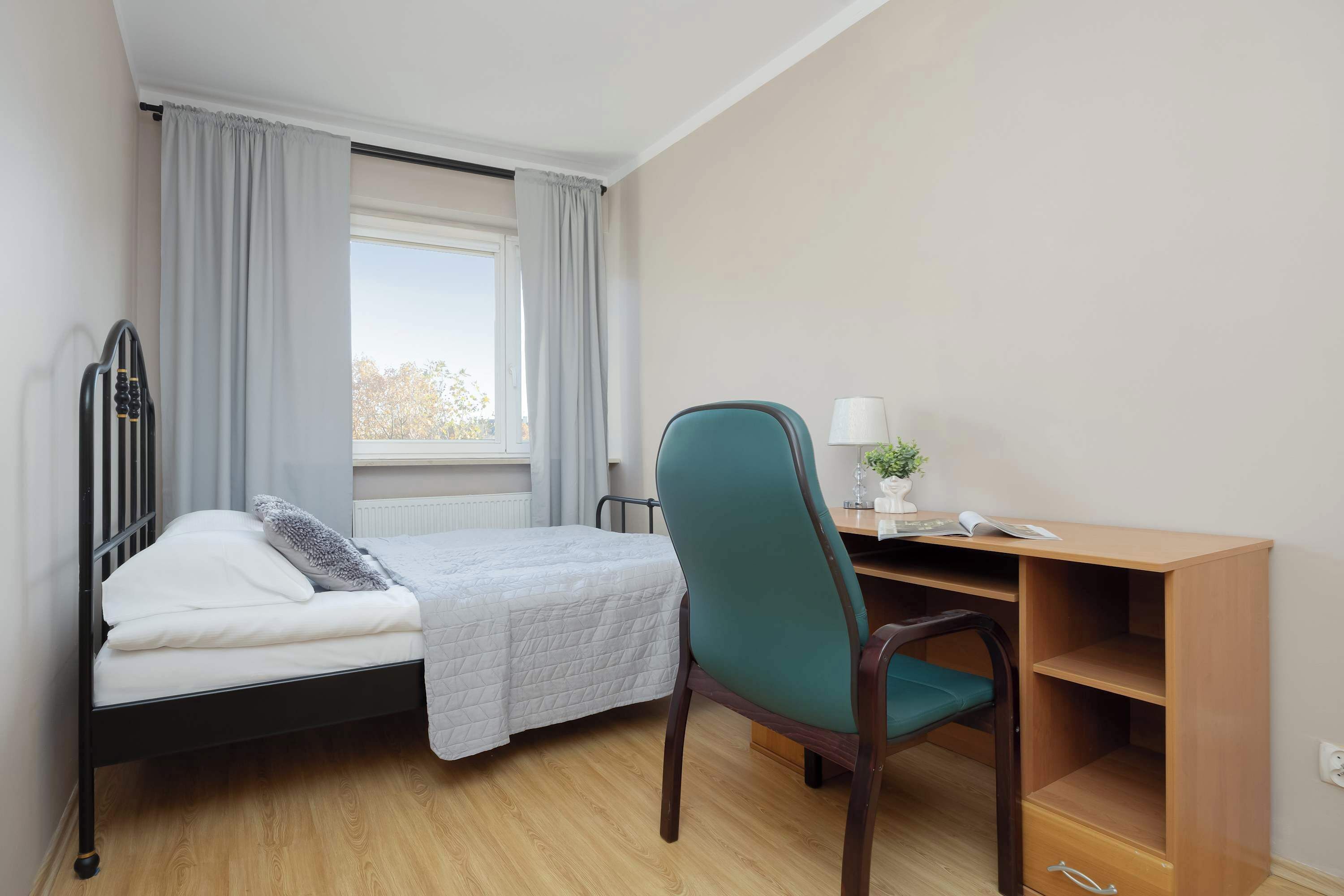 A bedroom with a bed, a desk, a chair, a window with curtains, and a radiator.
