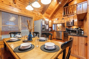 Interior de una cabaña de madera con una mesa de comedor preparada para cuatro personas, electrodomésticos de cocina y una zona de estar con sillas.