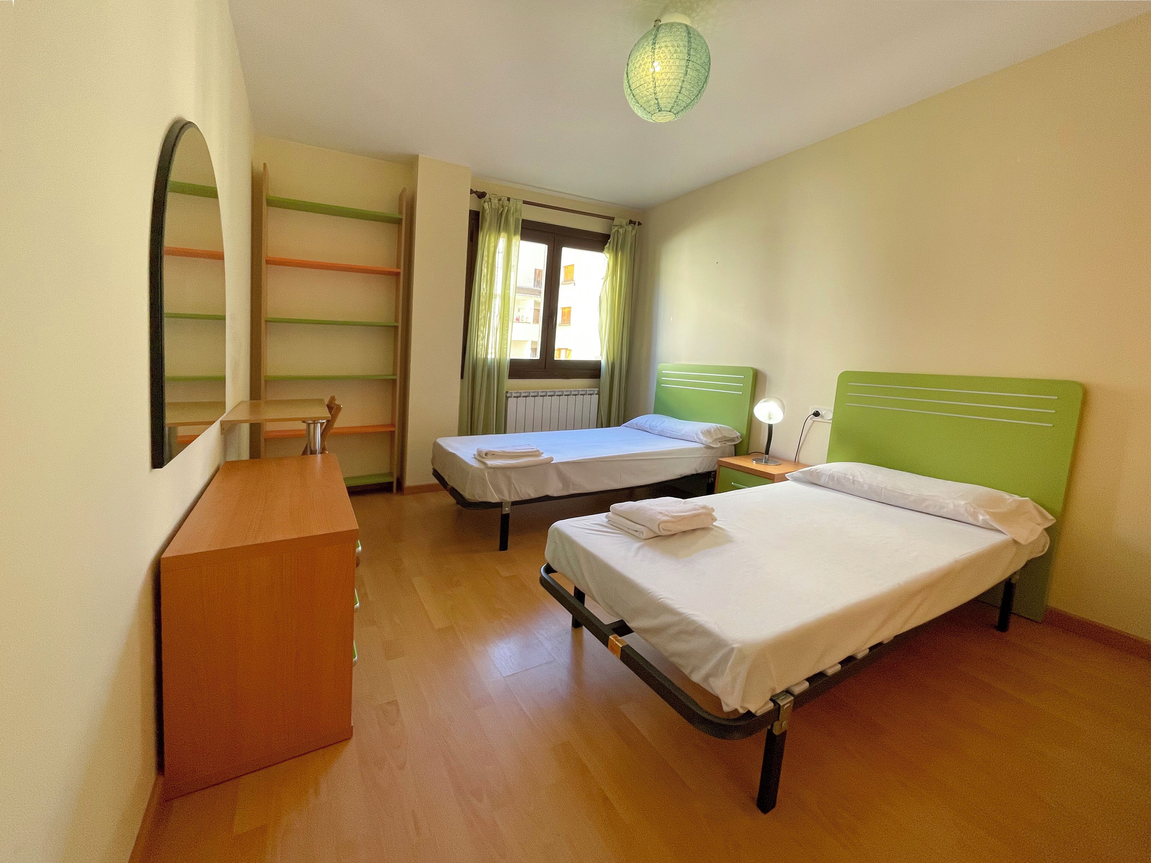 A hotel room with two single beds, a wooden desk, a mirror, and a green ceiling light.