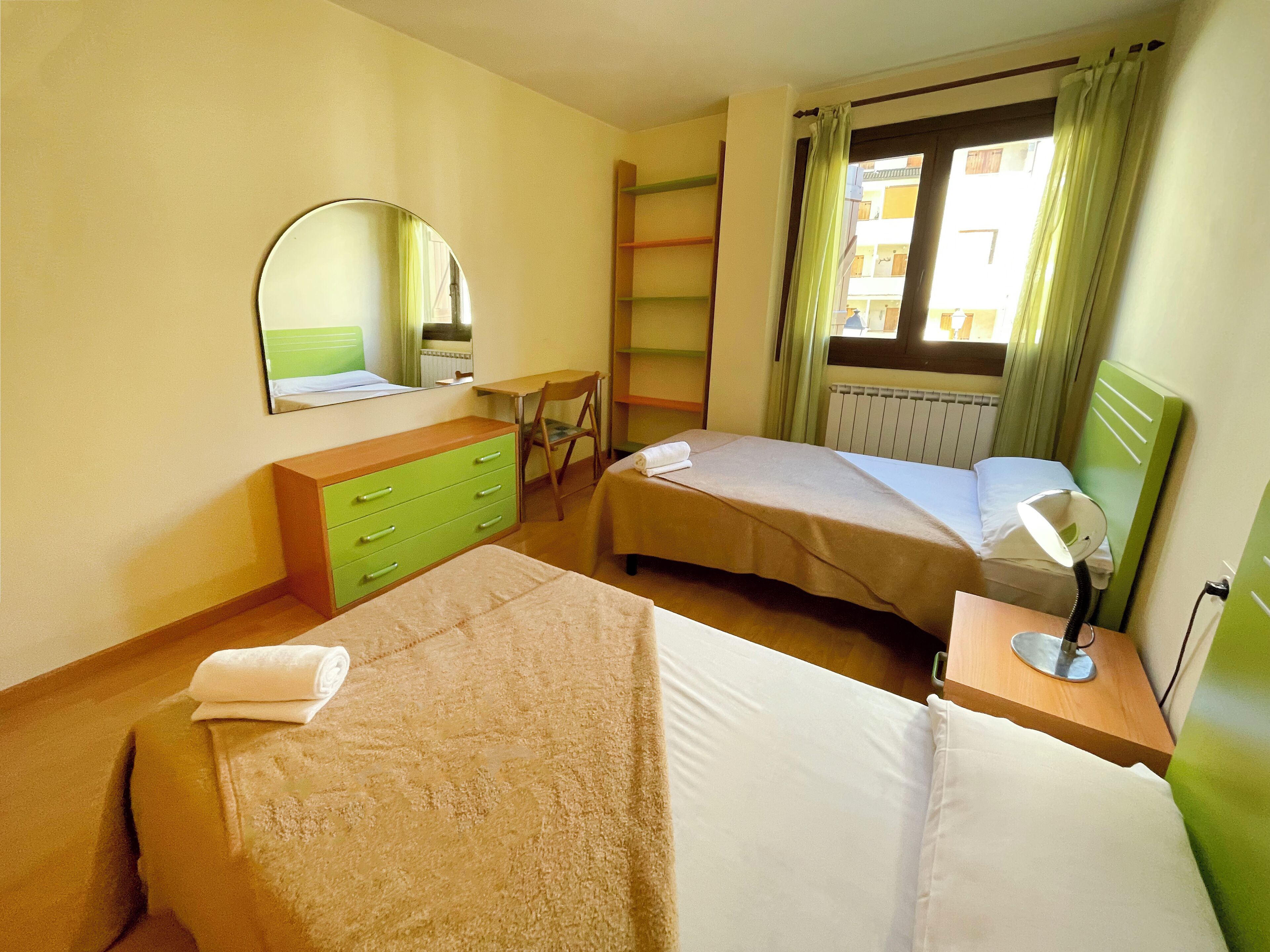 Habitación de hotel con dos camas, un escritorio, una silla, un espejo y un ventanal con cortinas.