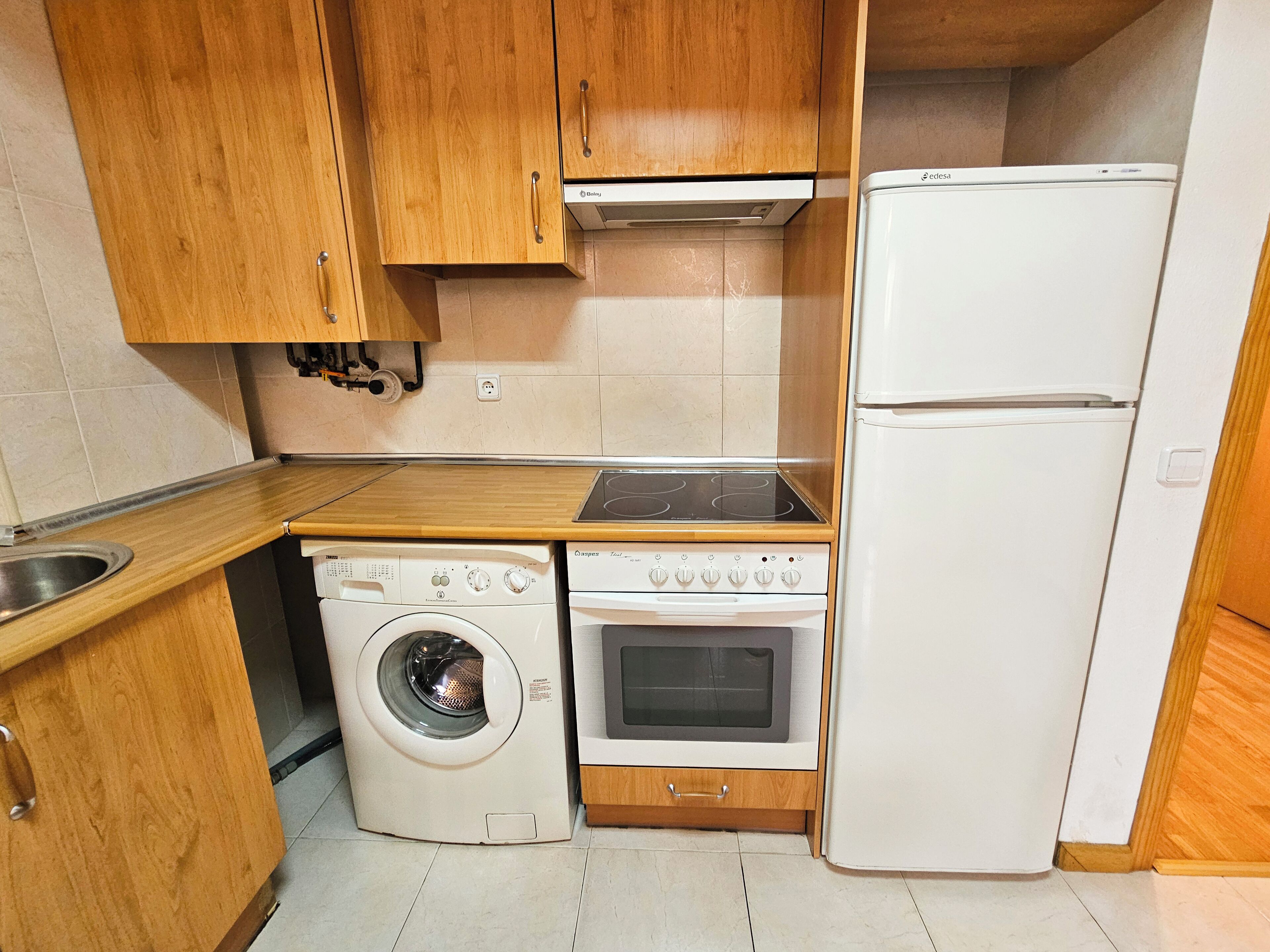Een kleine keuken met houten keukenkastjes, een ingebouwde wasmachine, een fornuis en een koelkast.