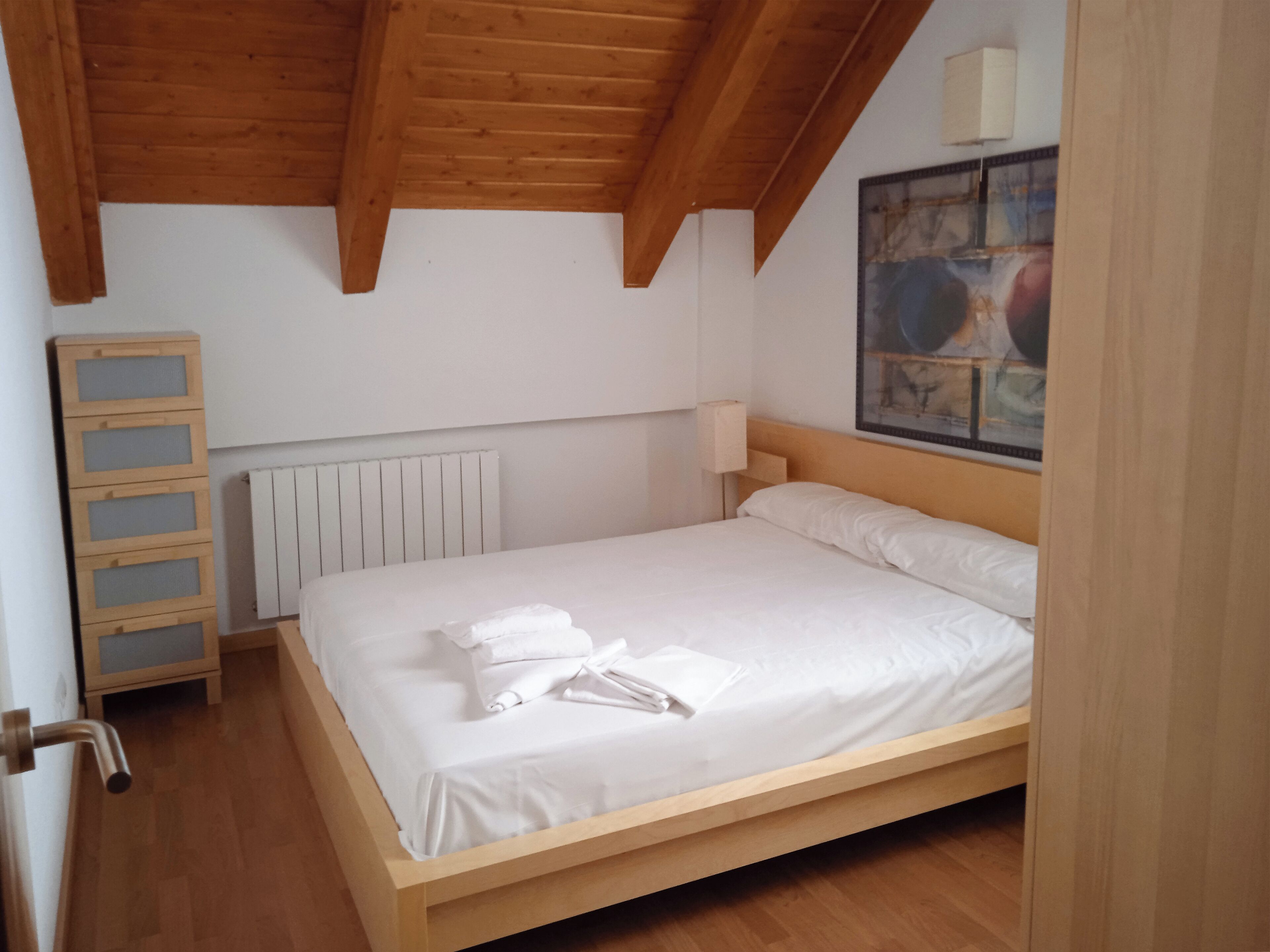 Un dormitorio con una cama de madera, ropa de cama blanca, un radiador, un armario de madera y un cuadro en la pared.