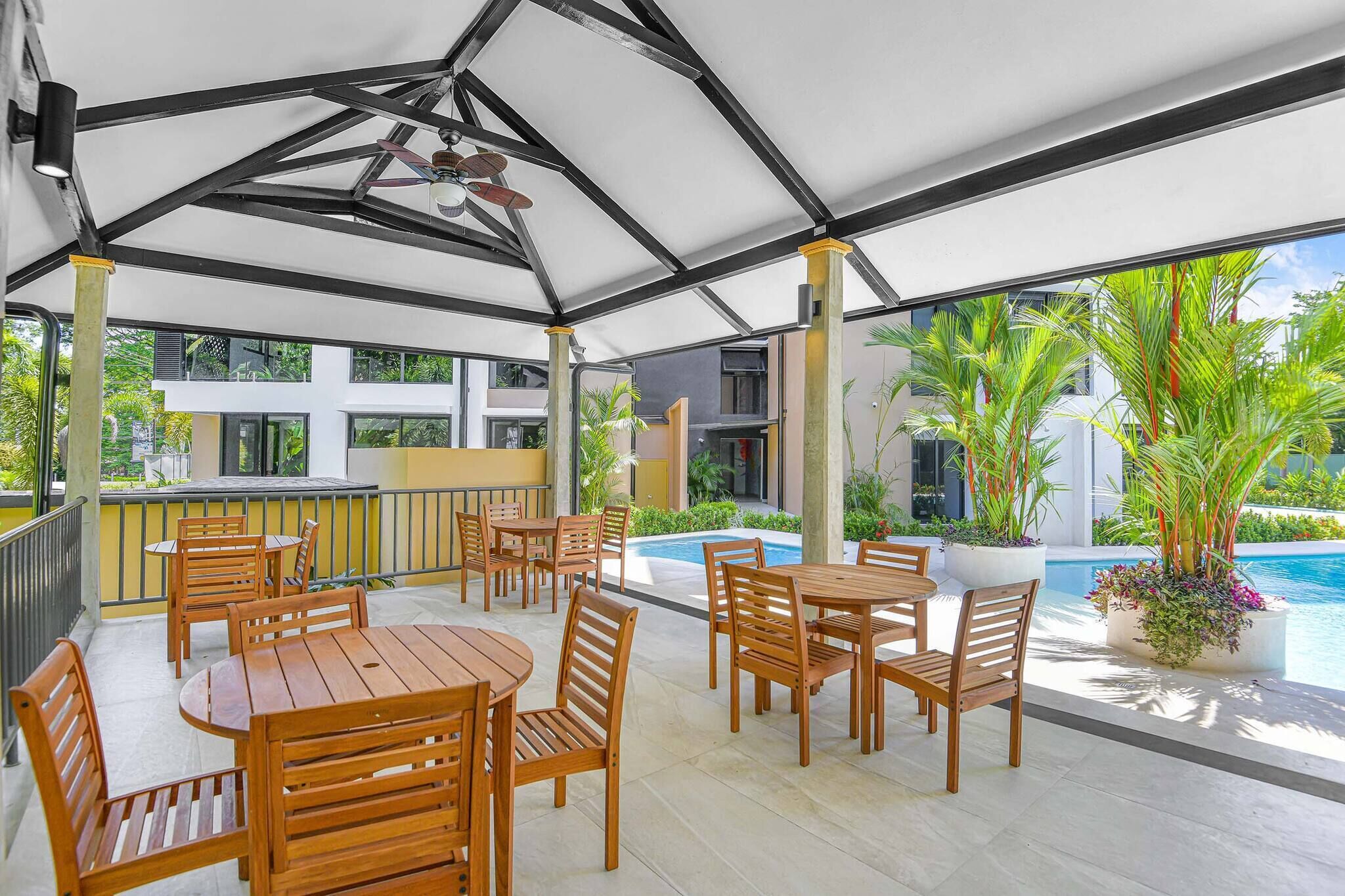 Ein überdachter Außenbereich mit Holztischen und -stühlen, einem Pool und Kübelpflanzen.