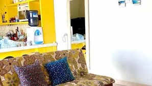 A living room with a sofa, a coffee table, and a kitchen area with yellow cabinets and a water dispenser. - Bale Jiwanmukti Narmada (Narmada)
