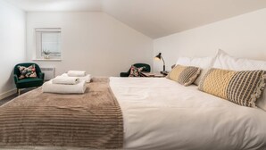 Een keurig opgemaakte slaapkamer met een groot bed, twee stoelen, een nachtkastje met een lamp en een raam met zonwering.