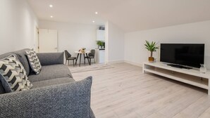 Een moderne woonkamer met een grijze bank, een flatscreen televisie op een witte kast, en een eethoek met een tafel en stoelen.