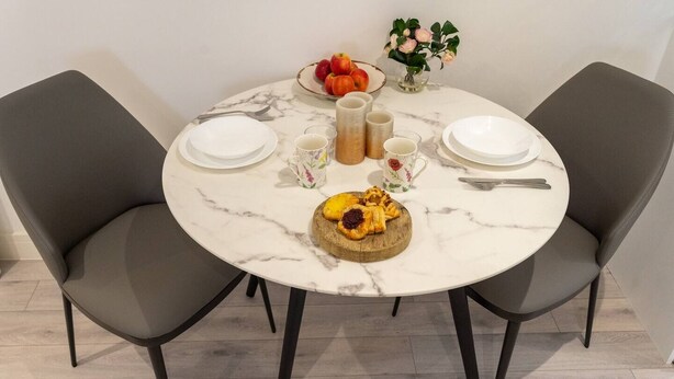 Een ronde marmeren tafel met twee grijze stoelen, een schaal met fruit, een vaas met bloemen en een bord gebak.