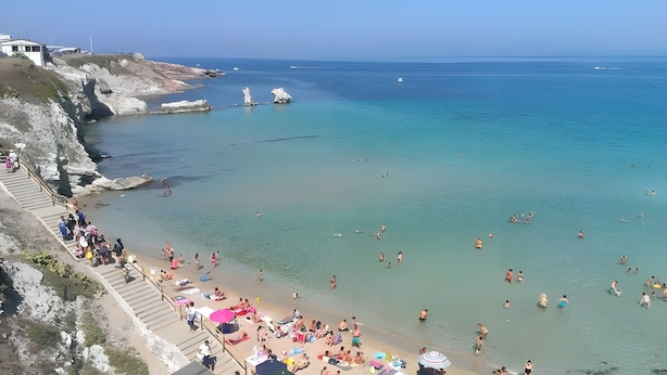 Ein Strand mit klarem Wasser, Menschen beim Schwimmen und Sonnenbaden sowie eine Treppe, die zum Ufer führt.