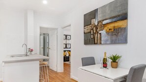A modern kitchen with a white countertop, bar stools, and a dining table with two chairs. A painting hangs on the wall above the table. - Ruthven Mansions CBD Boutique NY-Style Apartment (Adelaide)