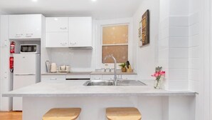 A modern kitchen with white cabinets, a built-in microwave, a sink, and two wooden stools. - Ruthven Mansions CBD Boutique NY-Style Apartment (Adelaide)