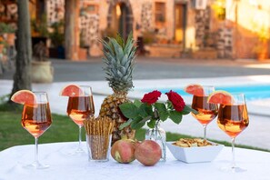 Uma mesa com bebidas, frutas e uma tigela de lanches, ao ar livre, com piscina e edifícios ao fundo.