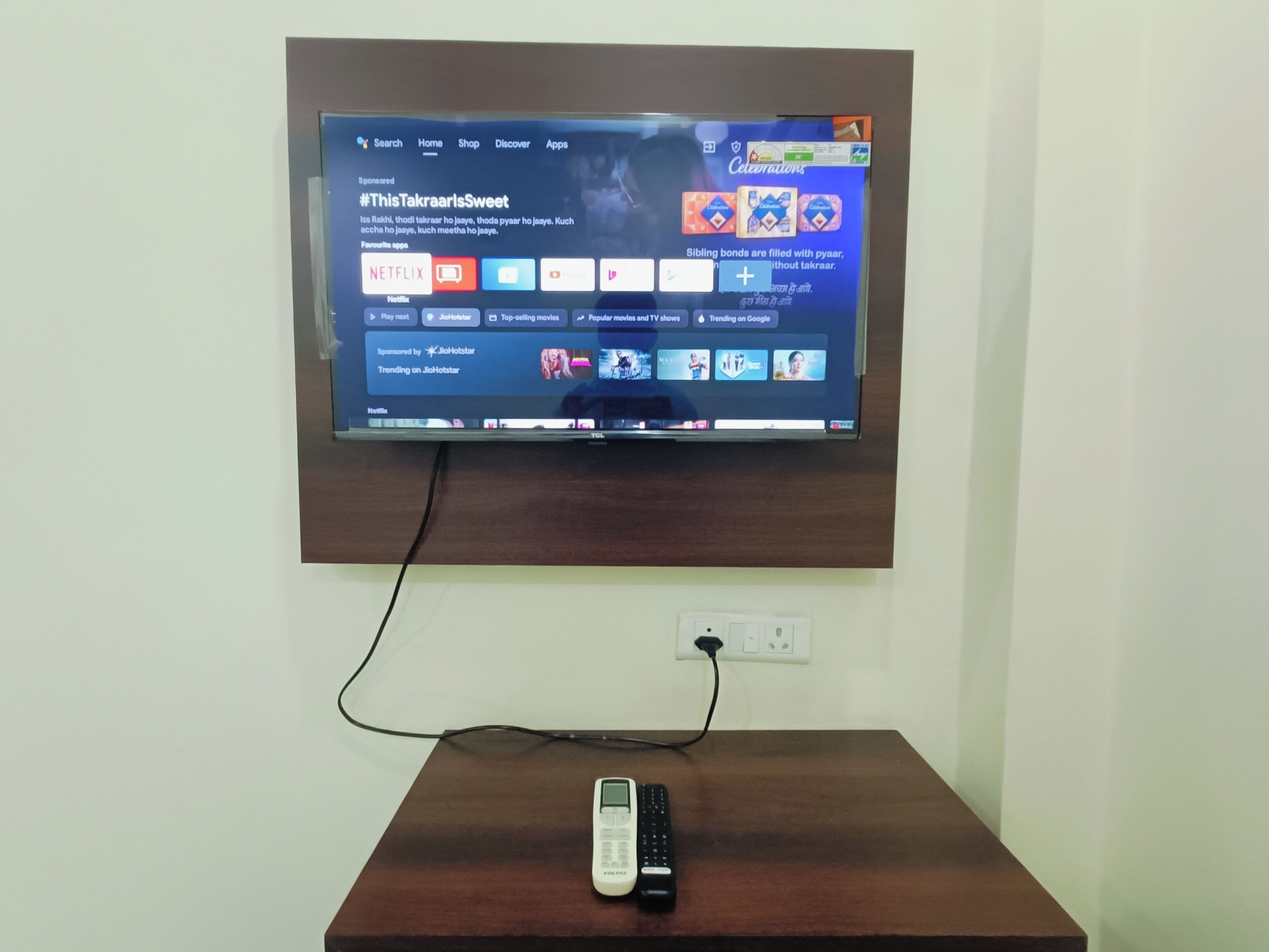 A flat-screen TV mounted on a wall above a wooden table with a remote control on it.