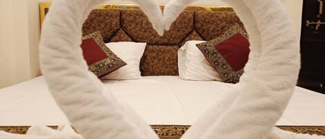 A bed with a heart-shaped towel arrangement, a patterned headboard, and a white wall with a light switch.