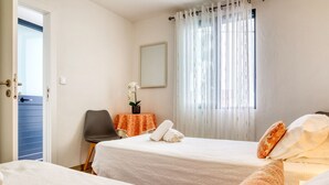 Une chambre d’hôtel avec deux lits, une chaise, une petite table ornée d’une orchidée et une fenêtre avec des rideaux.