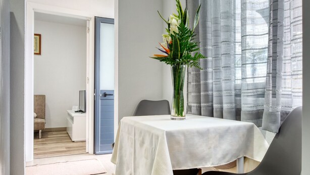 Un coin repas avec une table dressée pour deux personnes, un vase rempli de fleurs et une vue à travers des rideaux légers.