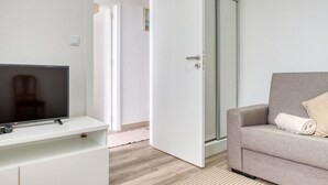 Une chambre moderne avec un téléviseur à écran plat posé sur un meuble blanc, un canapé gris avec une couverture enroulée, et une porte-fenêtre coulissante donnant sur une salle de bain.