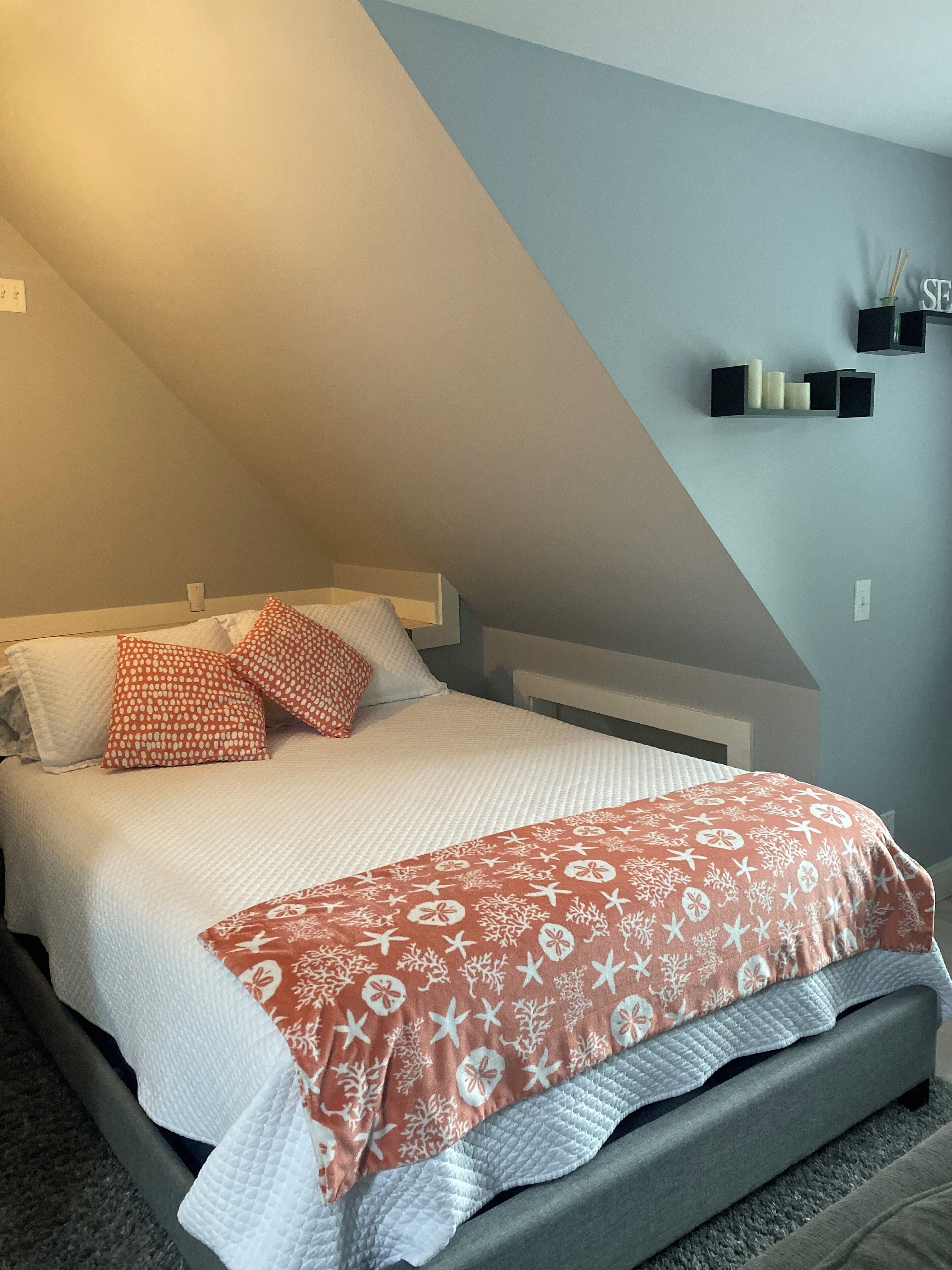 Une chambre à coucher avec un lit, des oreillers, une table de chevet et une étagère fixée au mur, sur laquelle sont posées des bougies.
